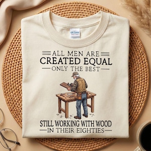 Puede incluir: Una camiseta de color crema con un gráfico de una persona trabajando en un banco de trabajo de madera con herramientas. El texto dice: ALL MEN ARE CREATED EQUAL ONLY THE BEST STILL WORKING WITH WOOD IN THEIR EIGHTIES. La camiseta tiene etiqueta Gildan.