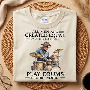 May include: A cream-coloured t-shirt featuring the text "ALL MEN ARE CREATED EQUAL - ONLY THE BEST STILL - PLAY DRUMS IN THEIR SEVENTIES." The graphic shows a drummer playing a drum kit.