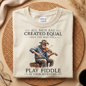 May include: A cream-coloured t-shirt with the text "ALL MEN ARE CREATED EQUAL - ONLY THE BEST STILL - PLAY FIDDLE IN THEIR SEVENTIES." The shirt features a watercolour illustration of a man playing the violin.