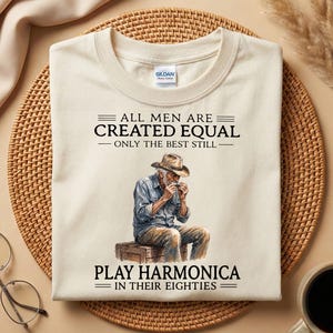 May include: A cream-colored short-sleeve t-shirt featuring a graphic of an elderly person in a hat playing a harmonica while sitting on a crate. The text reads: ALL MEN ARE CREATED EQUAL ONLY THE BEST STILL PLAY HARMONICA IN THEIR EIGHTIES.