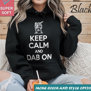 Peut inclure: Un sweat à capuche noir avec le texte "KEEP CALM AND DAB ON" en blanc. Le sweat à capuche présente un graphique d'une personne en train de faire un dab. Les mots "SUPER SOFT" et "Black" sont également visibles. Une citrouille avec un visage sculpté est au premier plan.