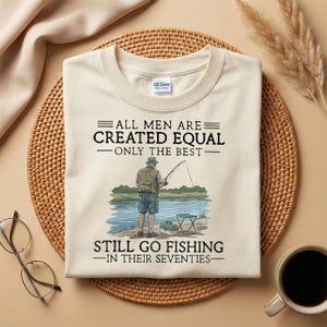 Könnte beinhalten: Cremefarbenes T-Shirt mit dem Text "ALL MEN ARE CREATED EQUAL - ONLY THE BEST - STILL GO FISHING - IN THEIR SEVENTIES." Eine Grafik zeigt einen Mann beim Angeln an einem See. Das Shirt liegt auf einer gewebten Matte.