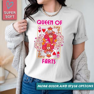 Op de afbeelding: Wit T-shirt met een speelkaartontwerp. De kaart toont een koningin in roze en oranje, omringd door roze wolken en harten, met de tekst "QUEEN OF FARTS". Het shirt heeft ook een "SUPER SOFT" logo.