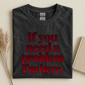 May include: A charcoal grey crew neck t-shirt with a vintage wash finish. The shirt features the red text "If you need a problem I'm here." printed in a bold serif font across the chest. A Gildan brand label is visible at the inner neckline.
