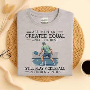 Könnte beinhalten: Ein hellgraues T-Shirt mit dem Text "ALLE MÄNNER SIND GLEICH GESCHAFFEN - NUR DIE BESTEN - SPIELEN NOCH PICKLEBALL - IN IHREN SIEBZIGERN." Eine Grafik zeigt eine Person beim Pickleballspielen. Das T-Shirt ist aus schwerer Baumwolle.