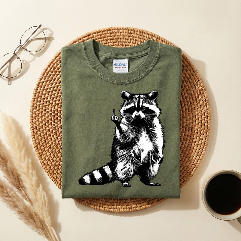 May include: Olive green t-shirt featuring a black and white illustration of a raccoon giving the middle finger. The raccoon is standing upright with its tail striped. The shirt has a crew neck and a Gildan label.