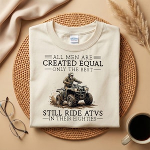 May include: A cream-coloured t-shirt with the text "ALL MEN ARE CREATED EQUAL - ONLY THE BEST - STILL RIDE ATVS - IN THEIR EIGHTIES." The graphic shows a person riding an ATV. The shirt is on a woven mat.