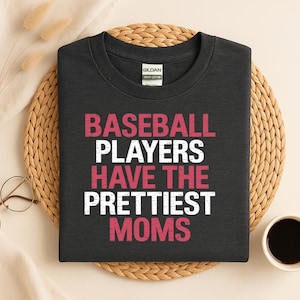 May include: Dark gray t-shirt with the text "BASEBALL PLAYERS HAVE THE PRETTIEST MOMS" in white and pink lettering. The shirt is made of heavy cotton and is displayed on a woven mat with a cup of coffee and eyeglasses.