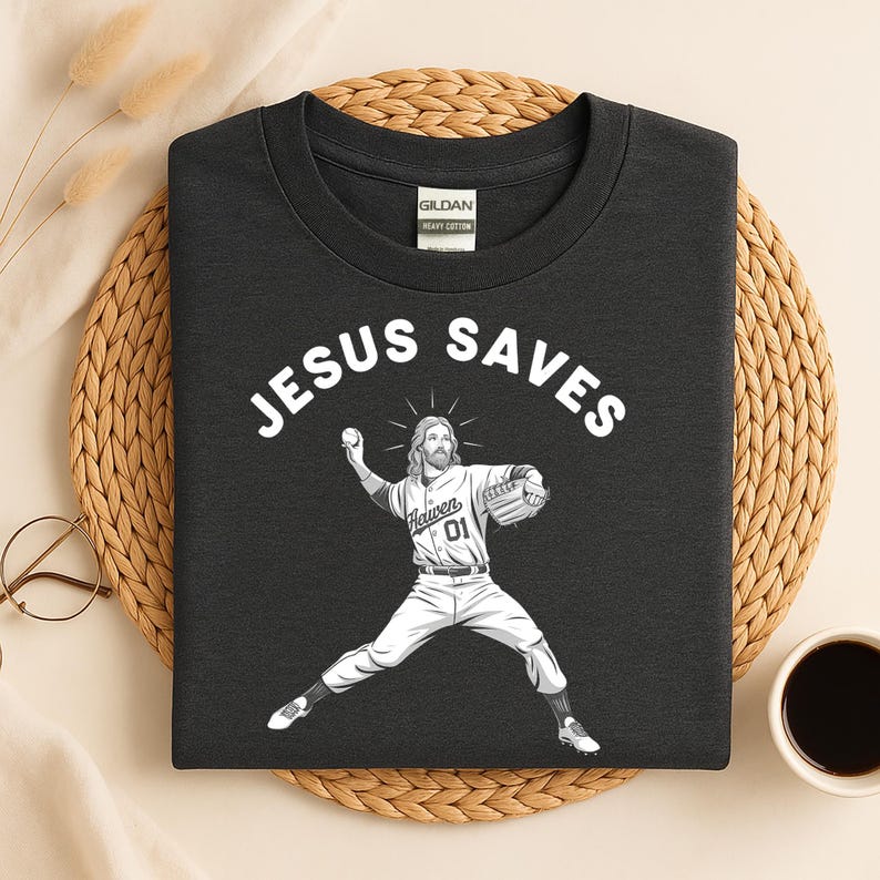 May include: Dark gray t-shirt with the text "JESUS SAVES" above an illustration of Jesus as a baseball pitcher. Jesus is wearing a baseball uniform with the word "Heaven" and the number "01" on the jersey.