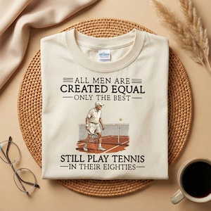 May include: Cream-colored t-shirt with the text "ALL MEN ARE CREATED EQUAL - ONLY THE BEST - STILL PLAY TENNIS - IN THEIR EIGHTIES." A graphic depicts a tennis player on a court. The shirt is on a woven mat with a coffee cup and glasses.