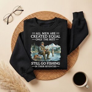 Op de afbeelding: Zwarte sweatshirt met de tekst "ALL MEN ARE CREATED EQUAL - ONLY THE BEST - STILL GO FISHING - IN THEIR SEVENTIES." De afbeelding toont een man die vist met een berg- en meerachtergrond.
