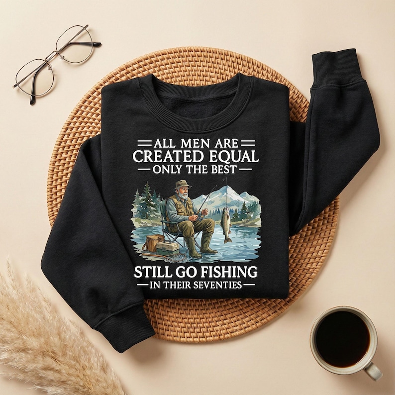 Op de afbeelding: Zwarte sweatshirt met de tekst "ALL MEN ARE CREATED EQUAL - ONLY THE BEST - STILL GO FISHING - IN THEIR SEVENTIES." De afbeelding toont een man die vist met een berg- en meerachtergrond.