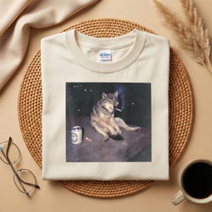 K&ouml;nnte beinhalten: Cremefarbenes T-Shirt mit einem Aufdruck eines Wolfs, der eine Zigarette raucht, mit einer Bierdose und einem Zigarettenstummel. Das Shirt hat einen Rundhalsausschnitt und den Markennamen "Gildan" auf dem Etikett.