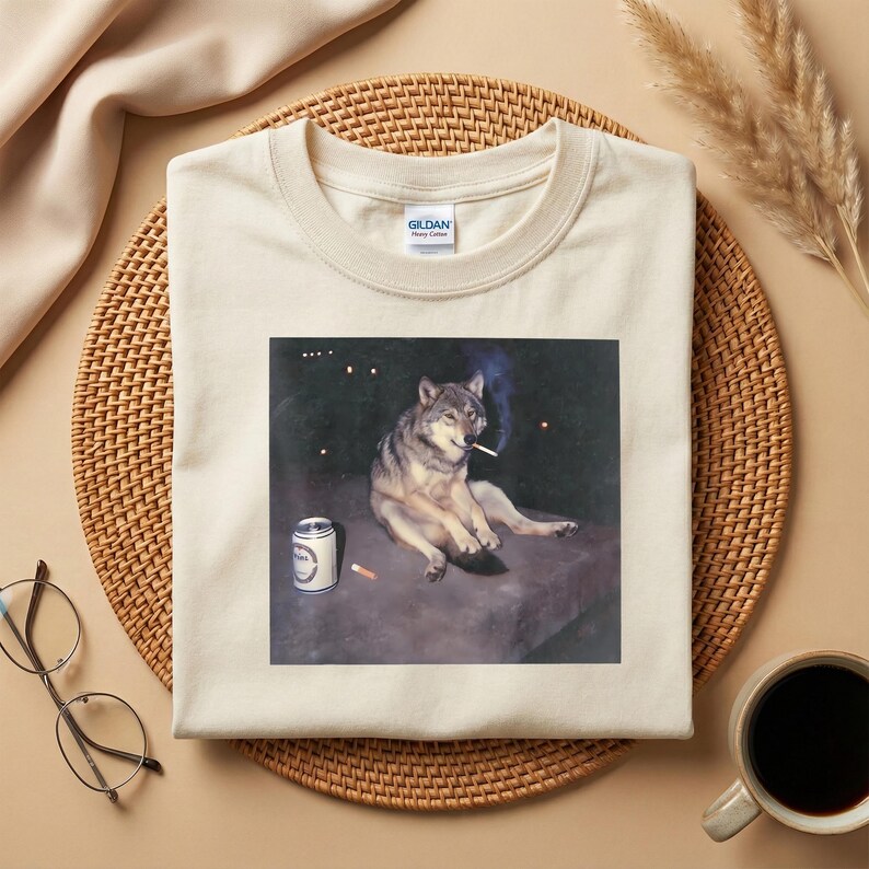 K&ouml;nnte beinhalten: Cremefarbenes T-Shirt mit einem Aufdruck eines Wolfs, der eine Zigarette raucht, mit einer Bierdose und einem Zigarettenstummel. Das Shirt hat einen Rundhalsausschnitt und den Markennamen "Gildan" auf dem Etikett.
