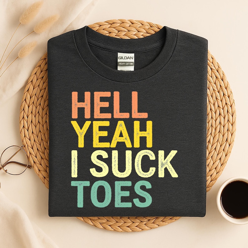Puede incluir: Una camiseta negra con la frase "HELL YEAH I SUCK TOES" en letras de colores. La camiseta est&aacute; hecha de algod&oacute;n grueso y tiene cuello redondo. La camiseta se muestra sobre una estera tejida.