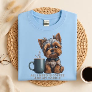 May include: Light blue t-shirt featuring a cartoon Yorkshire Terrier dog next to a coffee mug. The text "ALL I NEED IS COFFEE AND MY YORKIE" is printed below the image. The shirt is made of a soft, comfortable material.