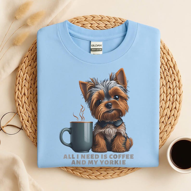 May include: Light blue t-shirt featuring a cartoon Yorkshire Terrier dog next to a coffee mug. The text "ALL I NEED IS COFFEE AND MY YORKIE" is printed below the image. The shirt is made of a soft, comfortable material.