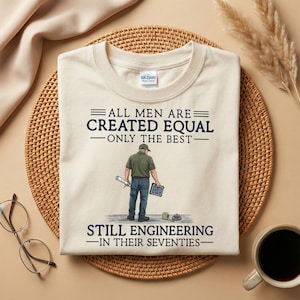 Puede incluir: Camiseta color crema con el texto "ALL MEN ARE CREATED EQUAL - ONLY THE BEST - STILL ENGINEERING - IN THEIR SEVENTIES." Un gráfico muestra a un ingeniero con planos y una tableta. La camiseta está sobre una estera tejida.