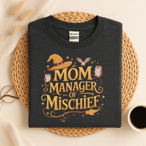 以下が含まれることがあります： 「MOM MANAGER OF MISCHIEF」の金色の文字が入った濃いグレーのTシャツ。デザインには魔女の帽子、フクロウ、星、ほうきが含まれています。このTシャツはヘビーコットン製です。