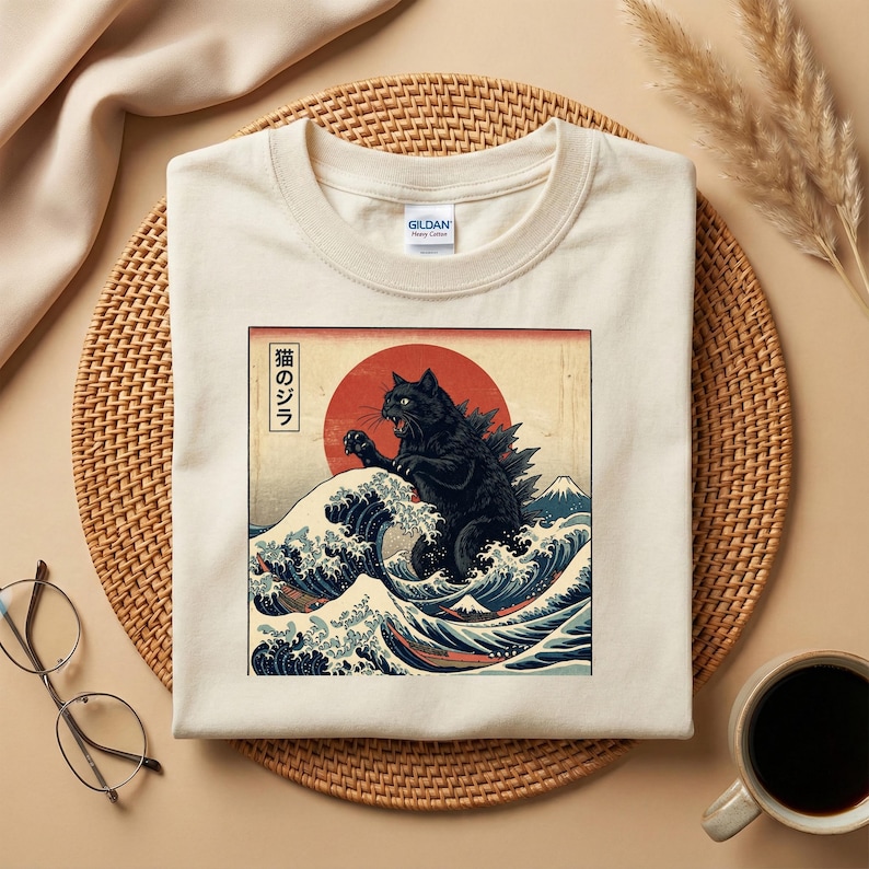 May include: Cream-colored t-shirt featuring a black cat-Godzilla hybrid design in a Japanese art style. The graphic includes a red sun, ocean waves, and Japanese text. The shirt is made of a soft material.