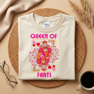 Op de afbeelding: Cr&egrave;mekleurig T-shirt met een speelkaartontwerp met een koningin, harten en de roze tekst "QUEEN OF FARTS". Het shirt wordt getoond op een geweven mat, met een koffiekopje en een bril in de buurt.