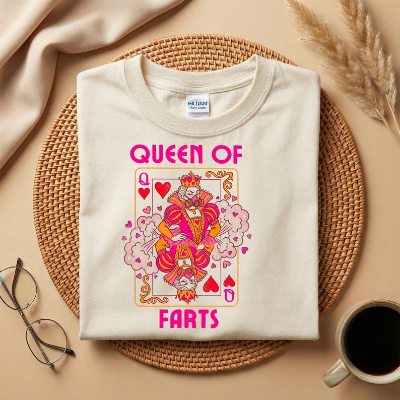Op de afbeelding: Cr&egrave;mekleurig T-shirt met een speelkaartontwerp met een koningin, harten en de roze tekst "QUEEN OF FARTS". Het shirt wordt getoond op een geweven mat, met een koffiekopje en een bril in de buurt.