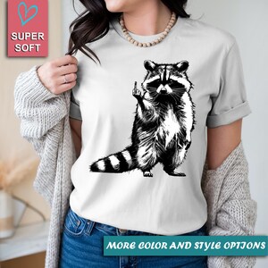 May include: White t-shirt featuring a black and white illustration of a raccoon giving the middle finger. The raccoon is standing upright with its tail striped. The words "SUPER SOFT" are in a pink box. The text "MORE COLOR AND STYLE OPTIONS" is at the bottom.