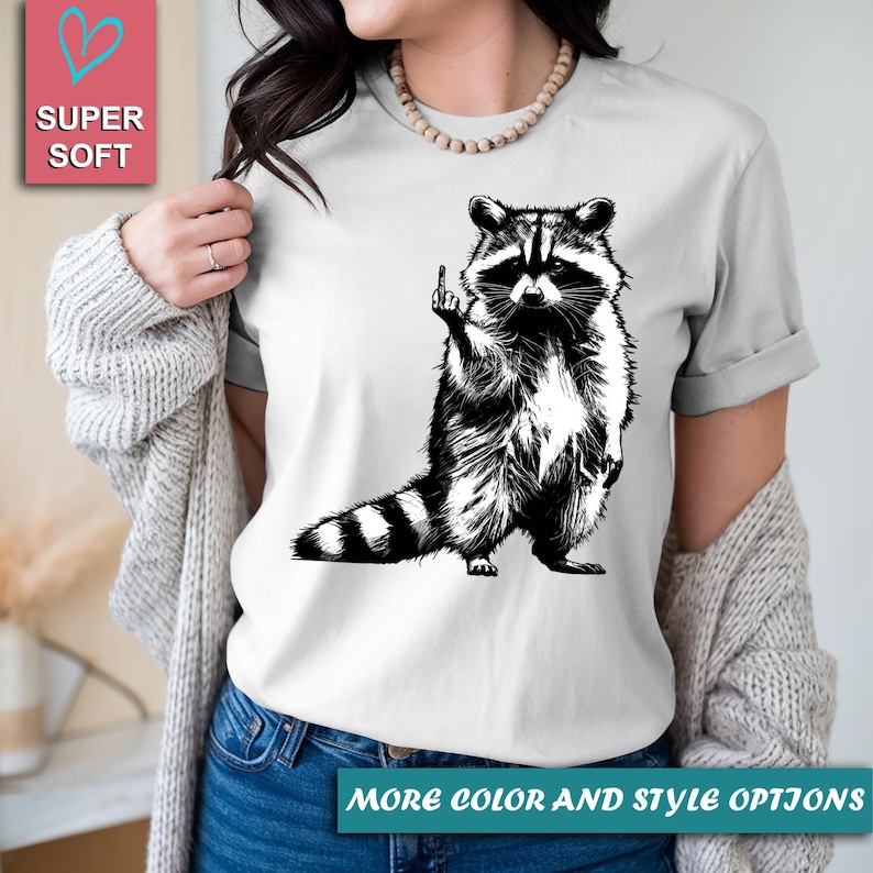 May include: White t-shirt featuring a black and white illustration of a raccoon giving the middle finger. The raccoon is standing upright with its tail striped. The words "SUPER SOFT" are in a pink box. The text "MORE COLOR AND STYLE OPTIONS" is at the bottom.