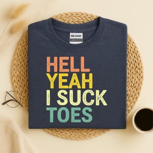 Puede incluir: Sudadera azul marino con el texto "HELL YEAH I SUCK TOES" en naranja, amarillo y turquesa. La sudadera es de algod&oacute;n grueso y se muestra sobre una esterilla tejida con una taza de caf&eacute; y gafas.