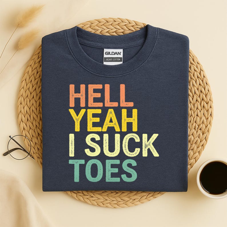Puede incluir: Sudadera azul marino con el texto "HELL YEAH I SUCK TOES" en naranja, amarillo y turquesa. La sudadera es de algod&oacute;n grueso y se muestra sobre una esterilla tejida con una taza de caf&eacute; y gafas.