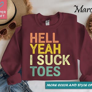 Puede incluir: Sudadera granate con el texto "HELL YEAH I SUCK TOES" en naranja, amarillo y turquesa. Las palabras est&aacute;n apiladas verticalmente. La imagen tambi&eacute;n incluye una etiqueta rosa en forma de coraz&oacute;n que dice "SUPER SOFT".