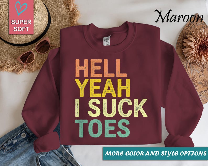 Puede incluir: Sudadera granate con el texto "HELL YEAH I SUCK TOES" en naranja, amarillo y turquesa. Las palabras est&aacute;n apiladas verticalmente. La imagen tambi&eacute;n incluye una etiqueta rosa en forma de coraz&oacute;n que dice "SUPER SOFT".
