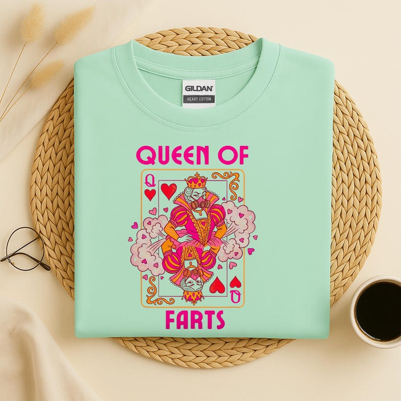 Op de afbeelding: Een lichtgroen t-shirt met de woorden "QUEEN OF FARTS" in roze. Het shirt heeft een afbeelding van een speelkaart met een koningin die een kroon en een gasmasker draagt. De kaart heeft harten en roze wolken.