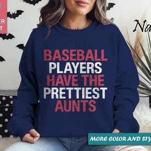 Op de afbeelding: Marineblauwe sweatshirt met de tekst "BASEBALL PLAYERS HAVE THE PRETTIEST AUNTS" in rood en wit. De sweatshirt is zacht en heeft een ronde hals. De afbeelding bevat ook een roze hartafbeelding en vleermuisdecoraties.