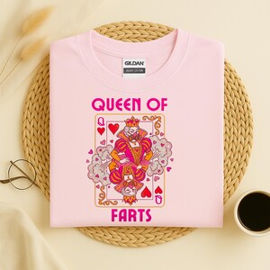 Op de afbeelding: Een lichtroze t-shirt met de tekst "QUEEN OF FARTS" in roze. De afbeelding is een speelkaartontwerp met een koningin die een kroon draagt en een roze en oranje outfit. Het shirt is gemaakt van zwaar katoen.