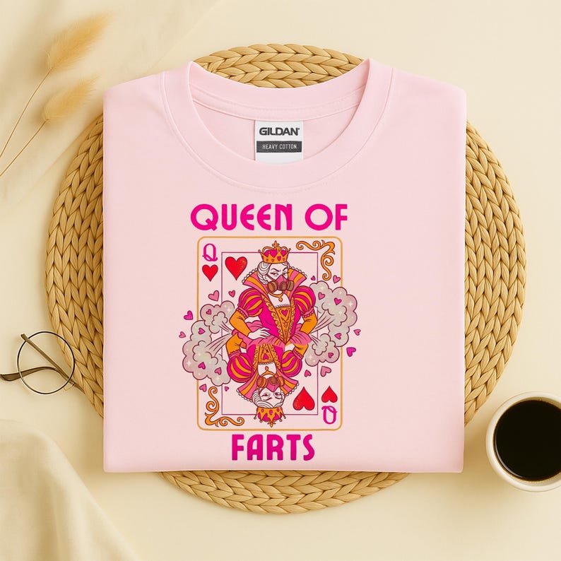 Op de afbeelding: Een lichtroze t-shirt met de tekst "QUEEN OF FARTS" in roze. De afbeelding is een speelkaartontwerp met een koningin die een kroon draagt en een roze en oranje outfit. Het shirt is gemaakt van zwaar katoen.