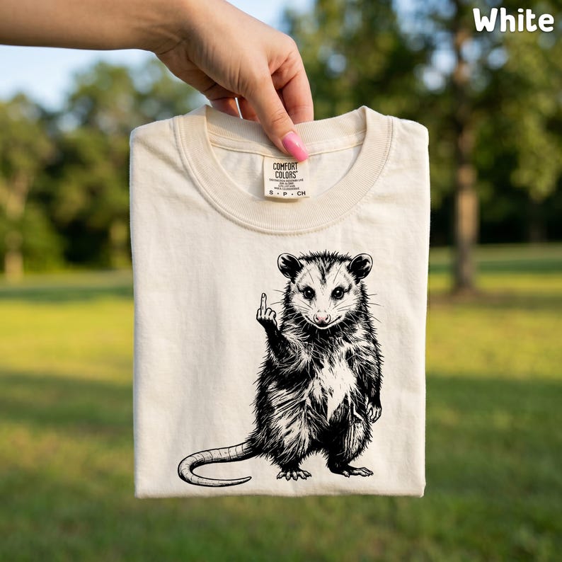 May include: Cream-colored t-shirt featuring a black and white illustration of an opossum giving the middle finger. The shirt is made of Comfort Colors material and has a crew neck. The word "White" is in the upper right corner.
