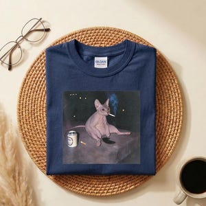 May include: A navy blue short-sleeve t-shirt featuring a graphic print of a hairless Sphynx cat sitting on a dark surface, holding a lit cigarette in its mouth with a small beverage can nearby. The shirt has a Gildan brand label at the neck.