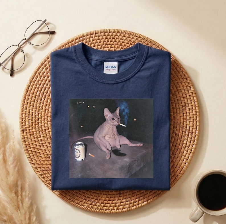 May include: A navy blue short-sleeve t-shirt featuring a graphic print of a hairless Sphynx cat sitting on a dark surface, holding a lit cigarette in its mouth with a small beverage can nearby. The shirt has a Gildan brand label at the neck.
