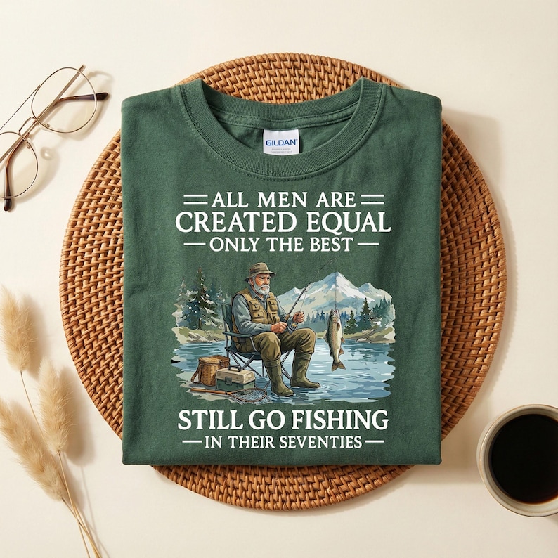 Op de afbeelding: Een donkergroen t-shirt met de tekst "ALL MEN ARE CREATED EQUAL - ONLY THE BEST - STILL GO FISHING - IN THEIR SEVENTIES." De afbeelding toont een oudere man die aan het vissen is. Het shirt is op een geweven mat gevouwen.