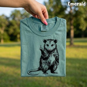 May include: Emerald green t-shirt featuring a black and white illustration of an opossum giving the middle finger. The shirt is folded and held up, with the brand label visible at the neckline. The word "Emerald" is printed in white in the top right corner.