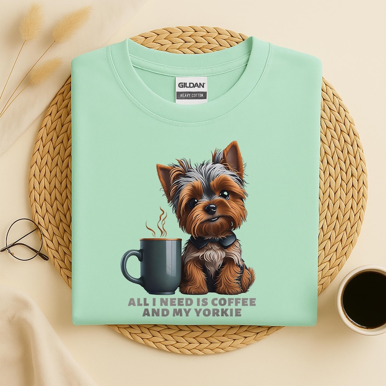 May include: A light green t-shirt featuring a cartoon Yorkshire Terrier next to a dark gray coffee mug with steam rising. The text "ALL I NEED IS COFFEE AND MY YORKIE" is printed below the mug and dog.