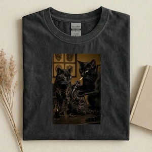 May include: Dark gray t-shirt featuring a graphic of two black cats, one tattooing the other. The tattooed cat has a spotted coat. A small bottle sits on the table. The shirt is made of heavy cotton.