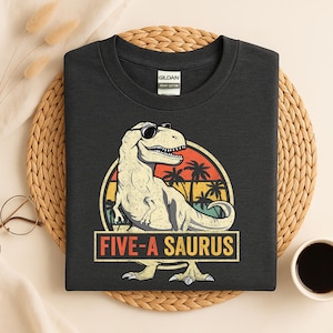 Op de afbeelding: Donkergrijs T-shirt met een dinosaurusafbeelding met zonnebril, tegen een zonsondergang met palmbomen. De tekst "FIVE-A SAURUS" staat eronder in een rechthoek. Het T-shirt is gemaakt van zwaar katoen.