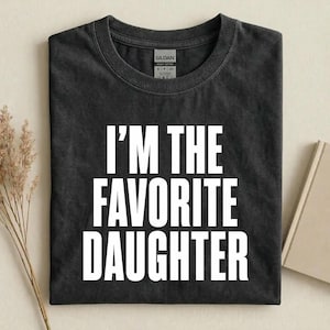 May include: A charcoal grey short-sleeved t-shirt with a crew neckline. The front features the white text "I'M THE FAVORITE DAUGHTER" in a bold, sans-serif font. The shirt has a Gildan brand label visible at the inner neck area.