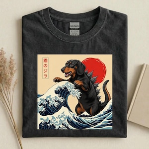 May include: A charcoal grey t-shirt featuring a graphic print of a black and tan dachshund standing in a large blue ocean wave. Behind the dog is a large red sun and Japanese text that reads 猫のジラ. The shirt has a Gildan brand label at the neck.