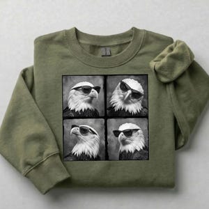 May include: An olive green sweatshirt featuring a black and white graphic of four eagles wearing sunglasses. The eagles are in a square grid pattern. The sweatshirt has a crew neck and long sleeves.