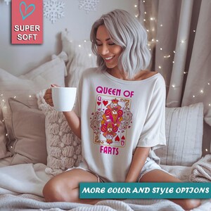 Op de afbeelding: Off-white t-shirt met een speelkaartafbeelding met de tekst "Queen of Farts". De afbeelding toont een cartoonkoningin en -koning met harten en kronen. Het shirt heeft ook de tekst "Super Soft" en "More Color and Style Options".