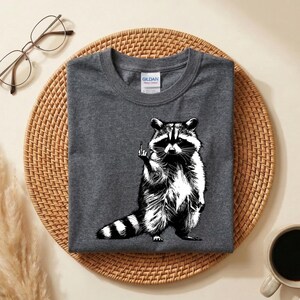 May include: Dark gray t-shirt featuring a black and white illustration of a raccoon giving the middle finger. The raccoon is standing upright with its tail striped. The shirt is folded on a woven mat.