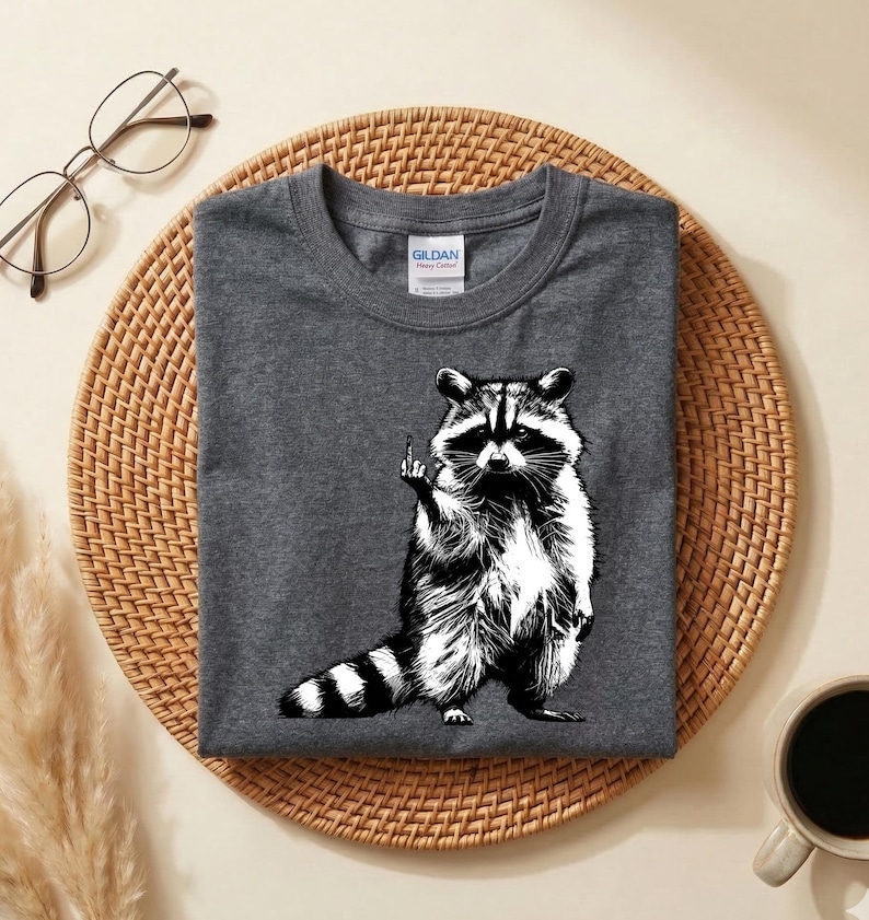 May include: Dark gray t-shirt featuring a black and white illustration of a raccoon giving the middle finger. The raccoon is standing upright with its tail striped. The shirt is folded on a woven mat.